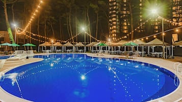 Una piscina con sillas de descanso y sombrillas, rodeada de edificios altos y luces colgantes.