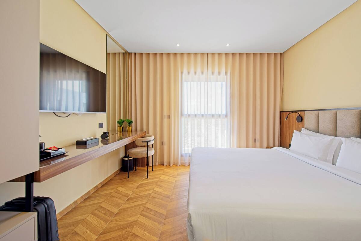 A hotel room with a large bed, a desk with a chair, a TV mounted on the wall, and a window with curtains.