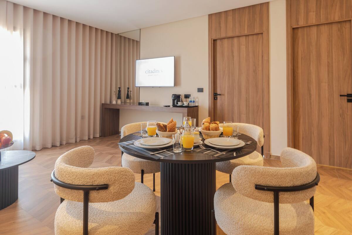 A modern dining area with a round table, four upholstered chairs, breakfast items, and a television on the wall.