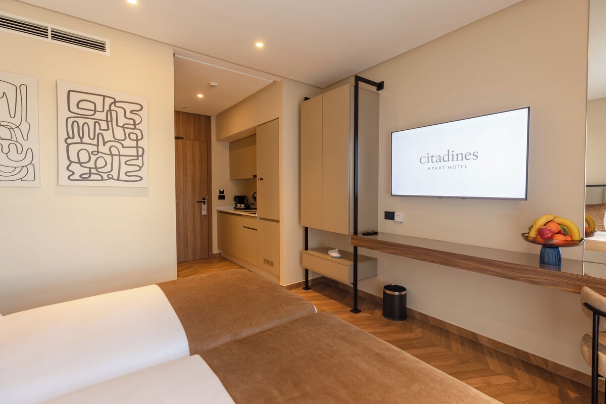 Una habitación de hotel moderna con una cama, un televisor, un frutero y una cocina visible a través de una puerta abierta.