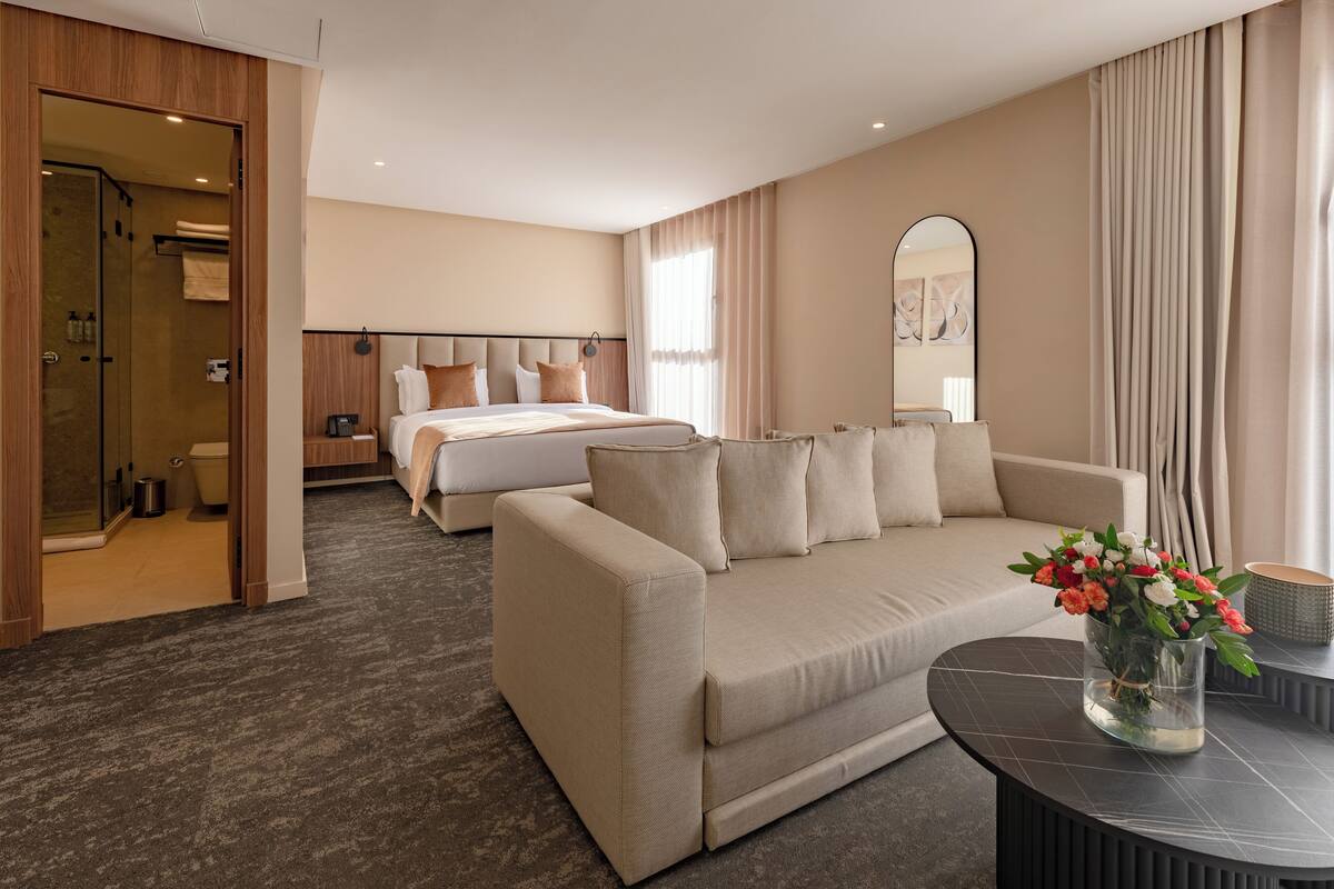 A hotel room with a bed, a sofa, a glass coffee table with flowers, and a shower area.