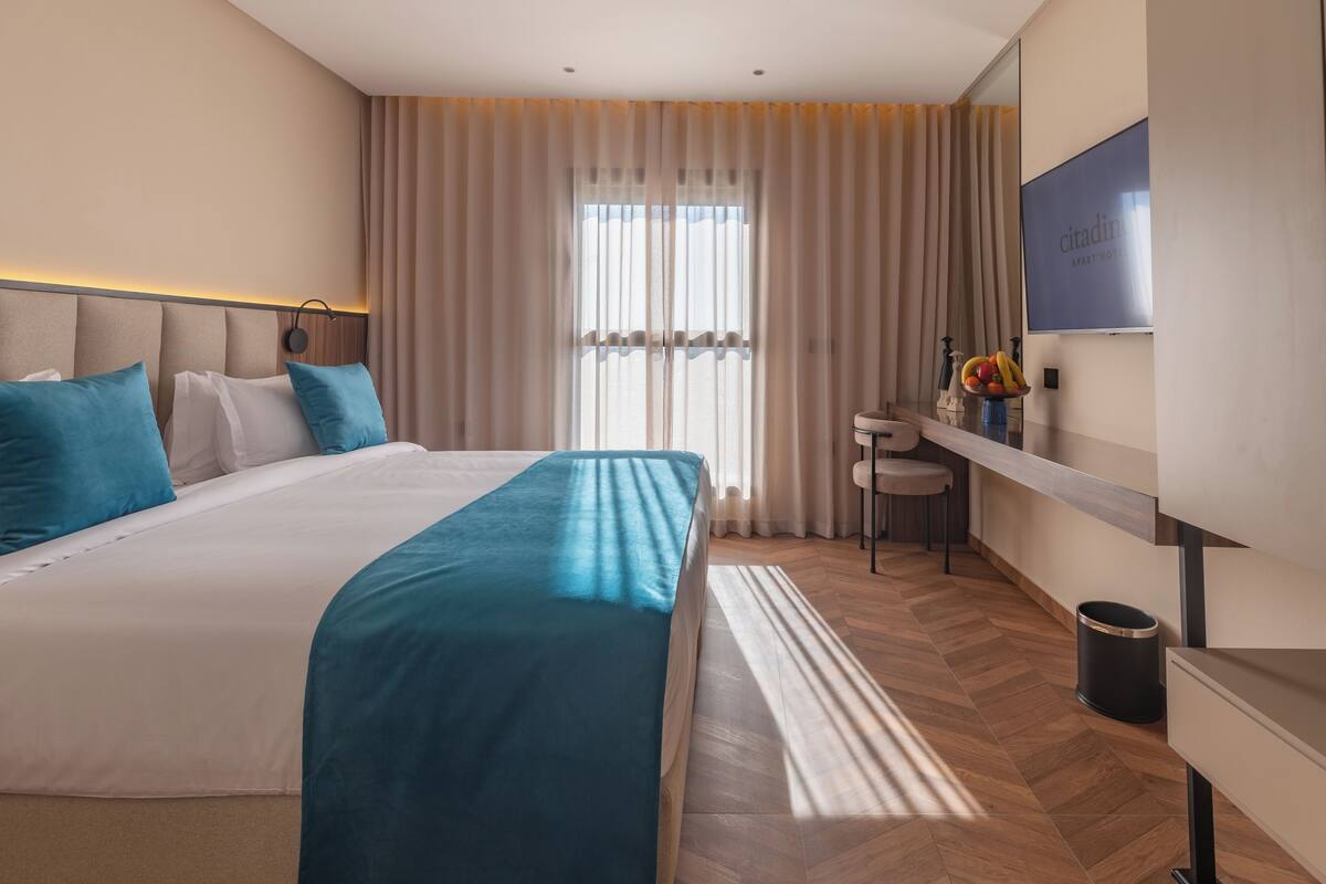 A hotel room with a large bed, a desk, a chair, a small table with a vase of fruit, and a television on the wall.