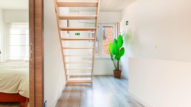 Ein modernes Hotelzimmer mit einer Holztreppe, einem Bett, einer Topfpflanze und einem Fenster mit Blick auf ein Ziegelgebäude.