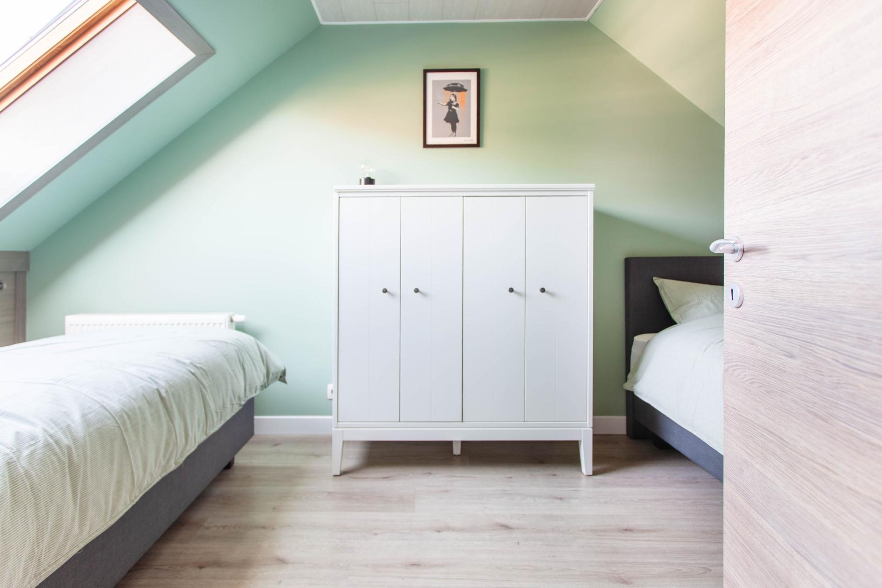 Une chambre avec un plafond en pente, une armoire blanche, deux lits et un tableau encadré au mur.
