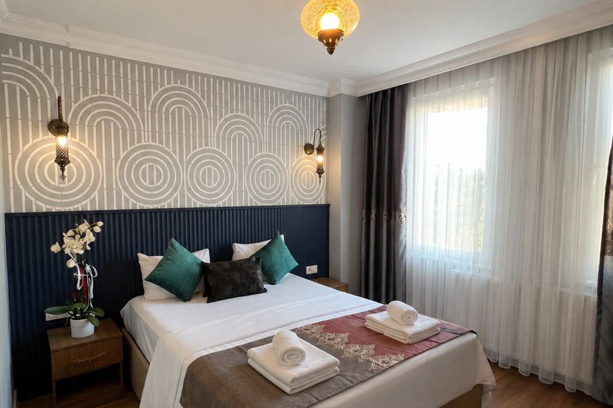 Habitación de hotel con una cama grande, una mesita de noche, una pared con diseño geométrico, una lámpara de techo y una ventana con cortinas.