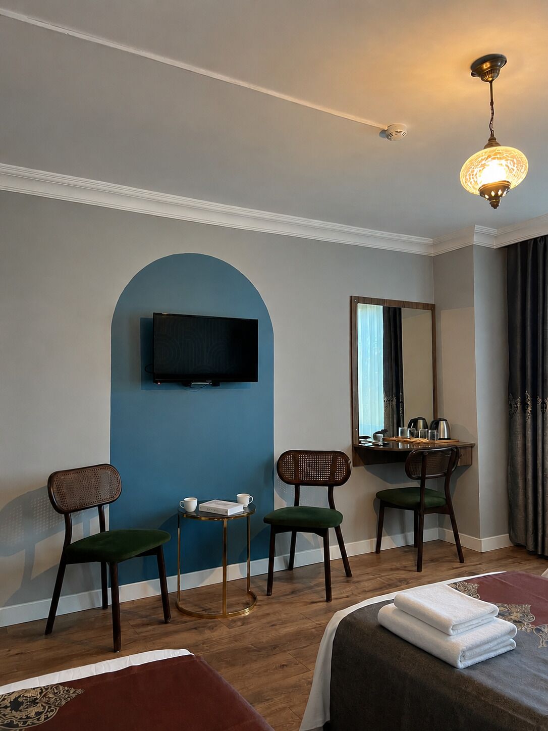 A hotel room with a television, two chairs, a small table, a bed with towels, a mirror, and a ceiling light.