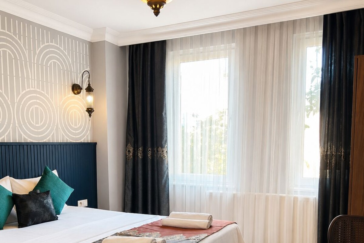 A hotel room with a bed, a desk, a chair, a hanging light fixture, and a window with curtains.