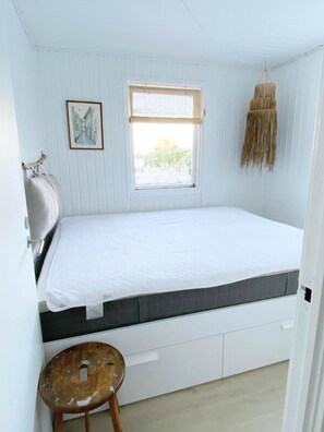 A small, white room with a bed, a window with a view, a picture on the wall, and a hanging macrame piece.