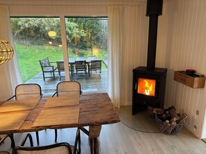 A wooden table with woven placemats, a wicker chair, a wooden bench, a fireplace with a visible fire, and a basket of firewood. - Lovely summer house with a fantastic view (Knebel)