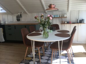 Et spiseområde med et rundt bord, brune stole, en vase med blomster og et køkken i baggrunden.
