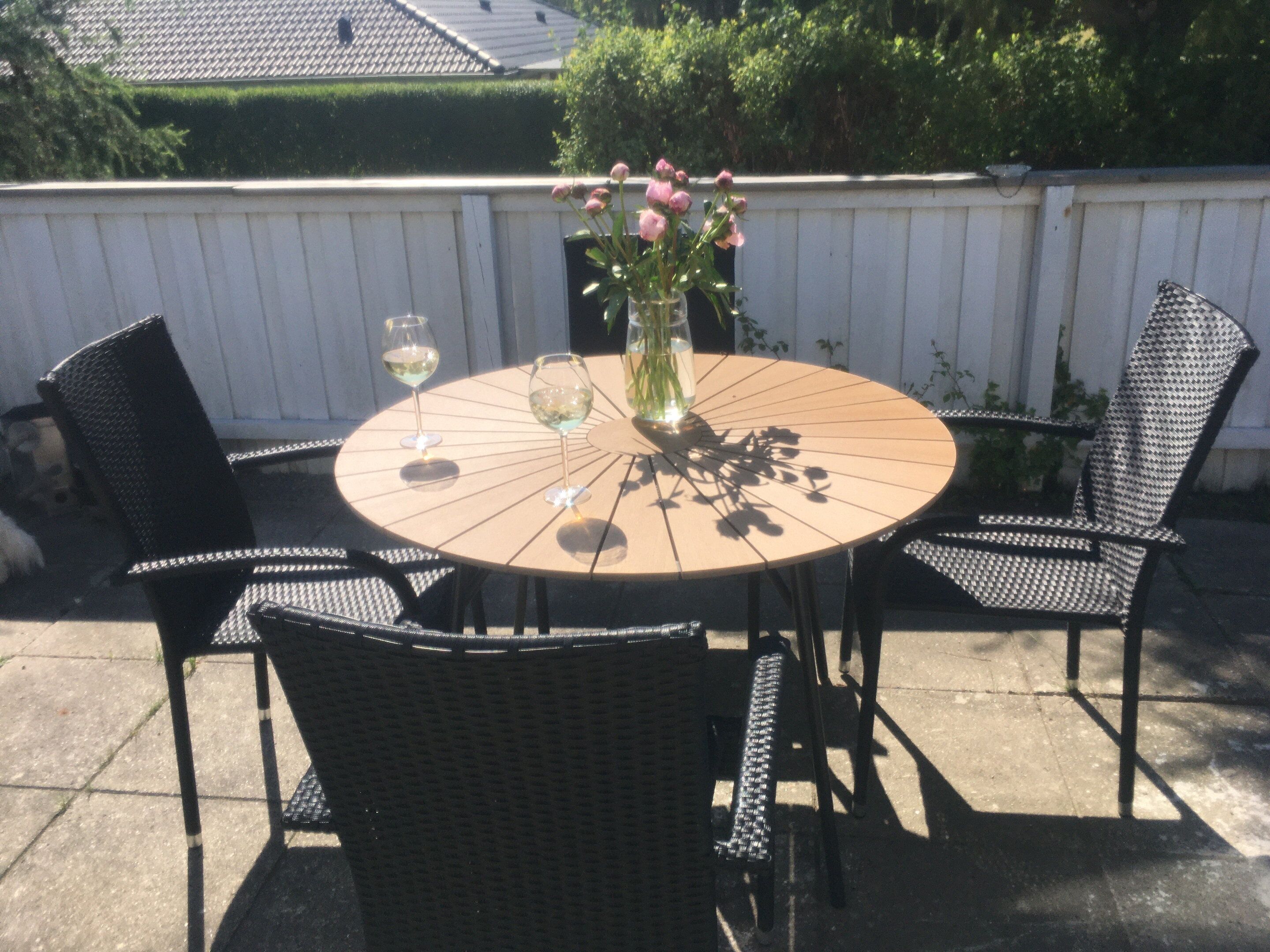 Ett runt utomhusbord med stolar, en vas med blommor och två vinglas.