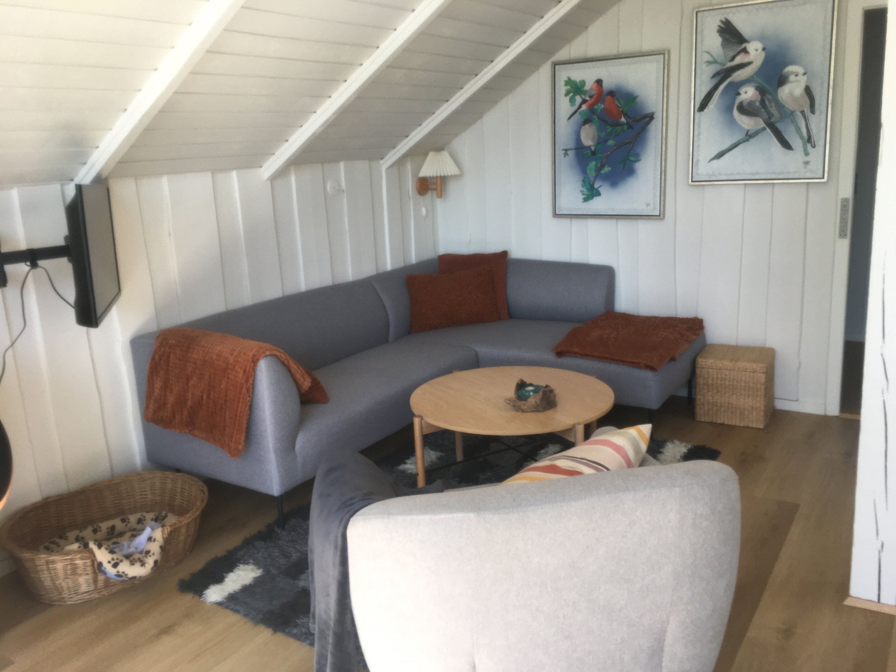 Un salon moderne avec un canapé d’angle gris, une table basse ronde en bois, un panier en osier et un tableau représentant un oiseau, accroché au mur.