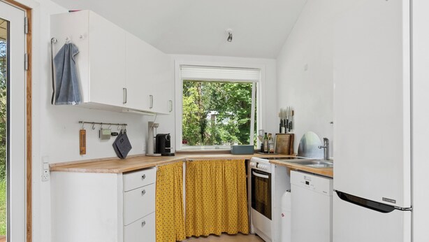 Een kleine keuken met witte kastjes, een raam en uitzicht op groen.