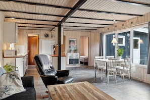 A modern interior with a wooden ceiling, a dining area with white chairs, a black leather sofa, and a kitchen area. - Cozy summer house near beach and nature (Væggerløse)