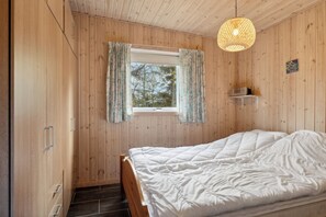 Une pièce aux murs lambrissés, avec un lit, une fenêtre avec des rideaux, une étagère contenant divers objets et un luminaire suspendu.