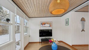A room with wooden floors, a large window, a black round table with a vase, a television, and a wall-mounted fish decoration. - Cozy beach bungalow close to Balka Beach (Nexø)