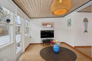 Una habitación con piso de madera, un ventanal grande, un televisor de pantalla plana y un jarrón azul con flores sobre una mesa redonda.