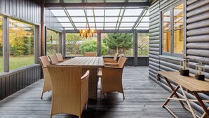 Un espacio comedor al aire libre techado, con una mesa de madera y sillas de mimbre, ventanas amplias y vistas de la naturaleza circundante.