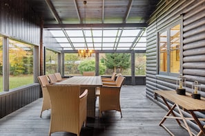 A covered outdoor dining area with a wooden table and wicker chairs, large windows, and a view of the surrounding nature.