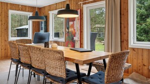 A wooden-paneled room with a dining table, wicker chairs, a pendant light, and large windows with curtains.