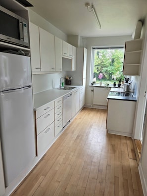 A kitchen with white cabinets, a built-in microwave, a refrigerator, a window with a view of greenery, and a countertop with a kettle.
