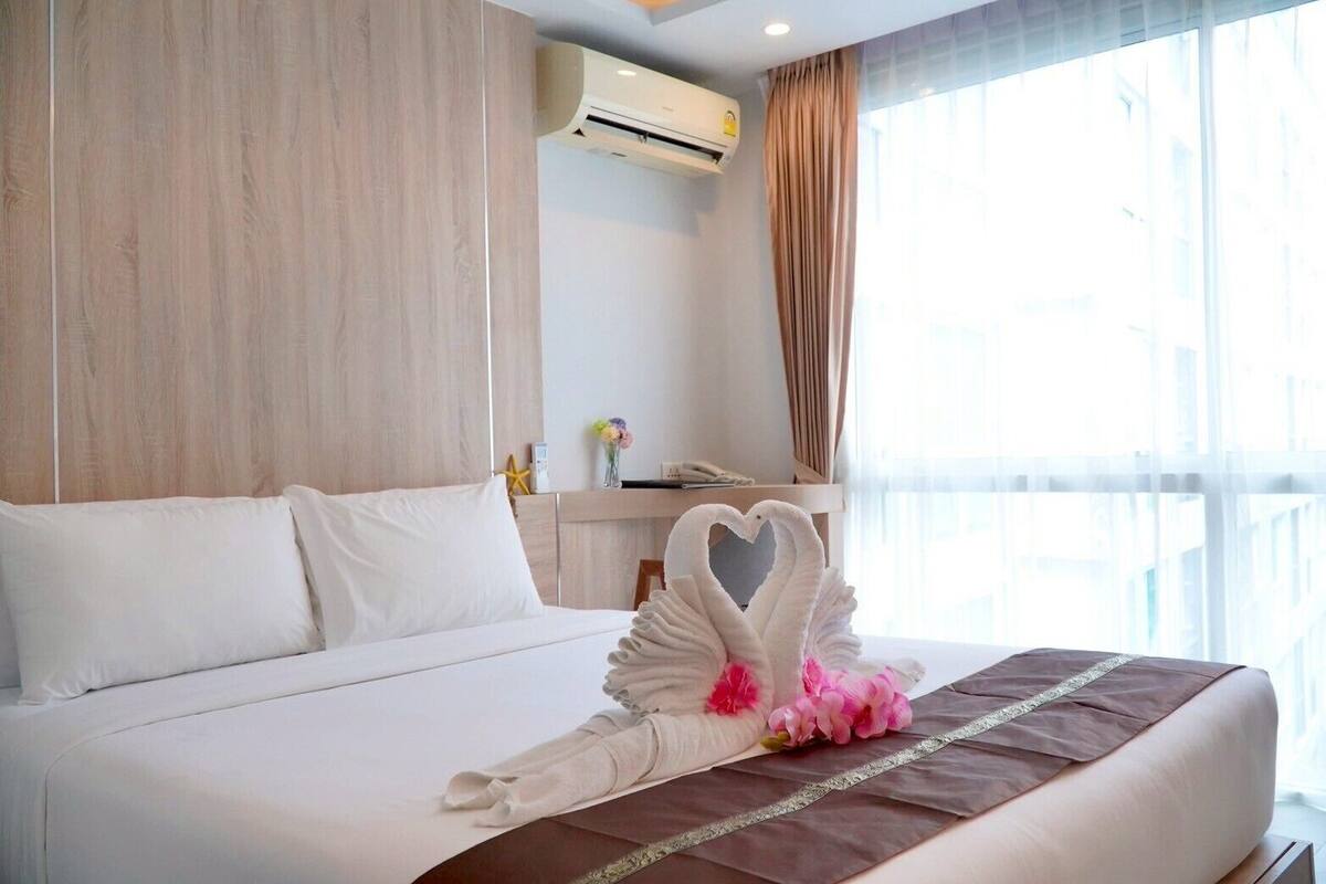 A hotel room with a large bed, a wooden headboard, an air conditioning unit, a desk with a phone, and a window with curtains.