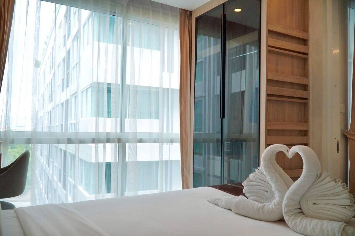 A hotel room with a large window, a bed with a heart-shaped towel, and a wooden wardrobe.