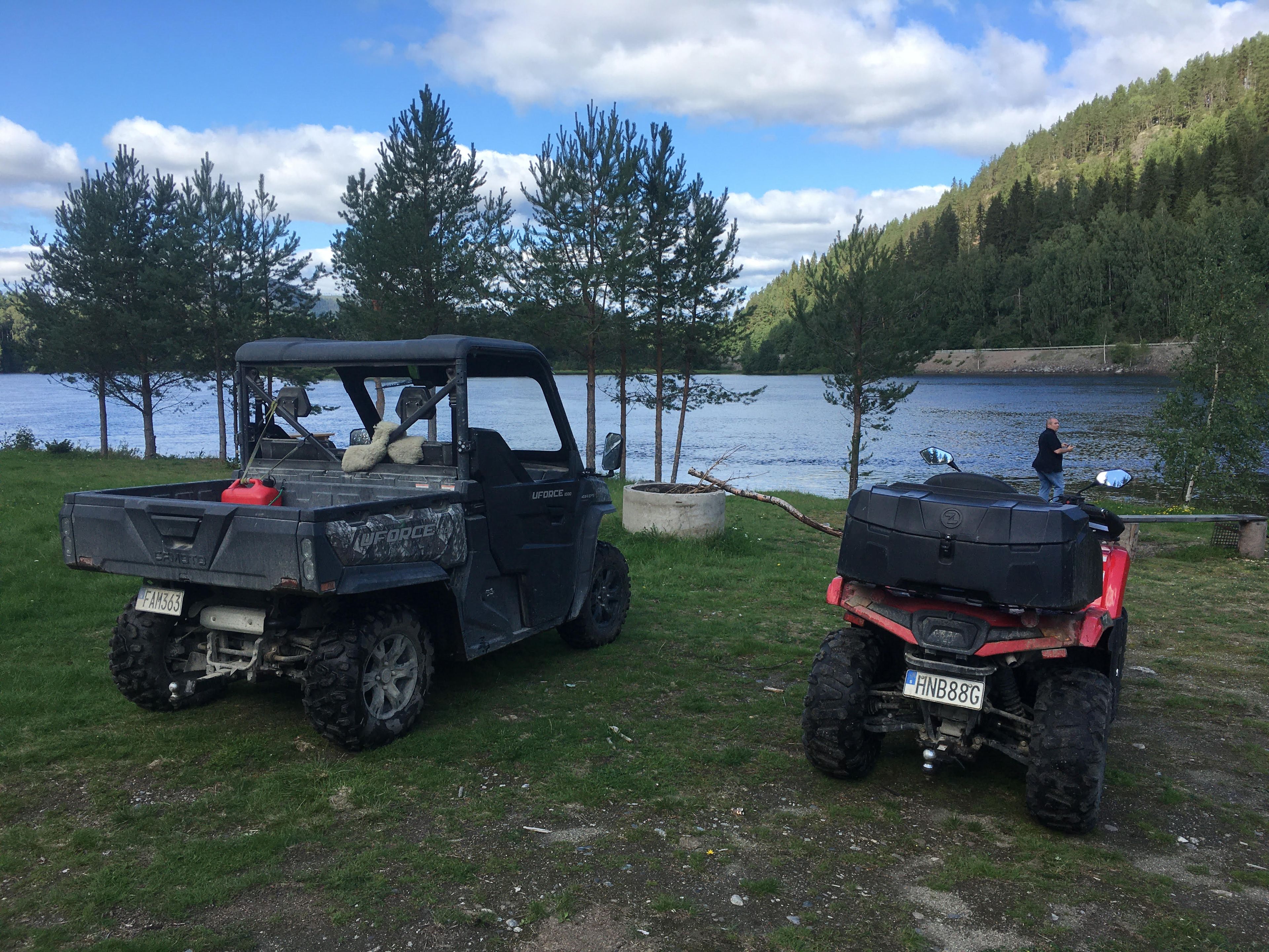Deux véhicules tout-terrain sont stationnés sur une zone herbeuse près d’un lac, avec des arbres et des collines en arrière-plan.