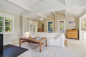 A room with a bed, a wooden table with books and a lamp, a dresser, and a view of greenery outside.