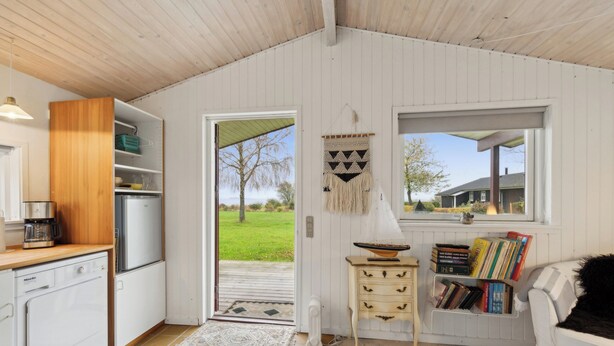 A compact kitchen with a refrigerator, coffee maker, and a wooden cabinet. A small table with books and a bowl is near a window. - Cozy little annex with a sea view. (Kalvehave)