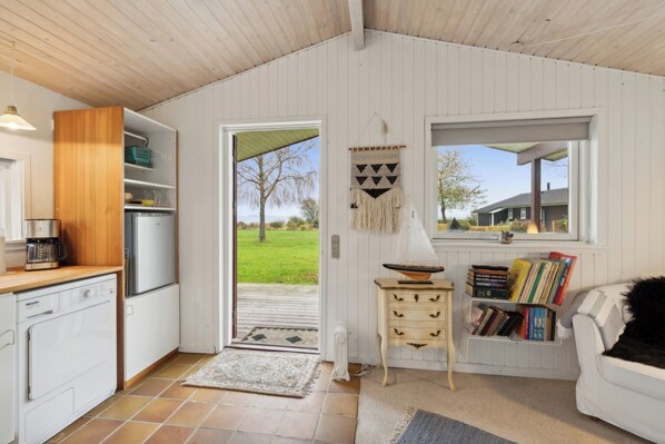 A compact kitchen with a refrigerator, coffee maker, and a wooden cabinet. A small table with books and a bowl is near a window.