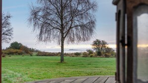 A view from a window overlooking a grassy field with a tree and distant horizon. - Cozy little annex with a sea view. (Kalvehave)