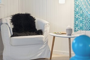 A white armchair with a black furry throw, a small round table with a white candle, and a blue object in the foreground.