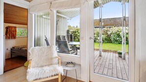 Une pièce avec un fauteuil blanc à la texture douce, une petite table et une vue sur une terrasse en bois et un jardin.
