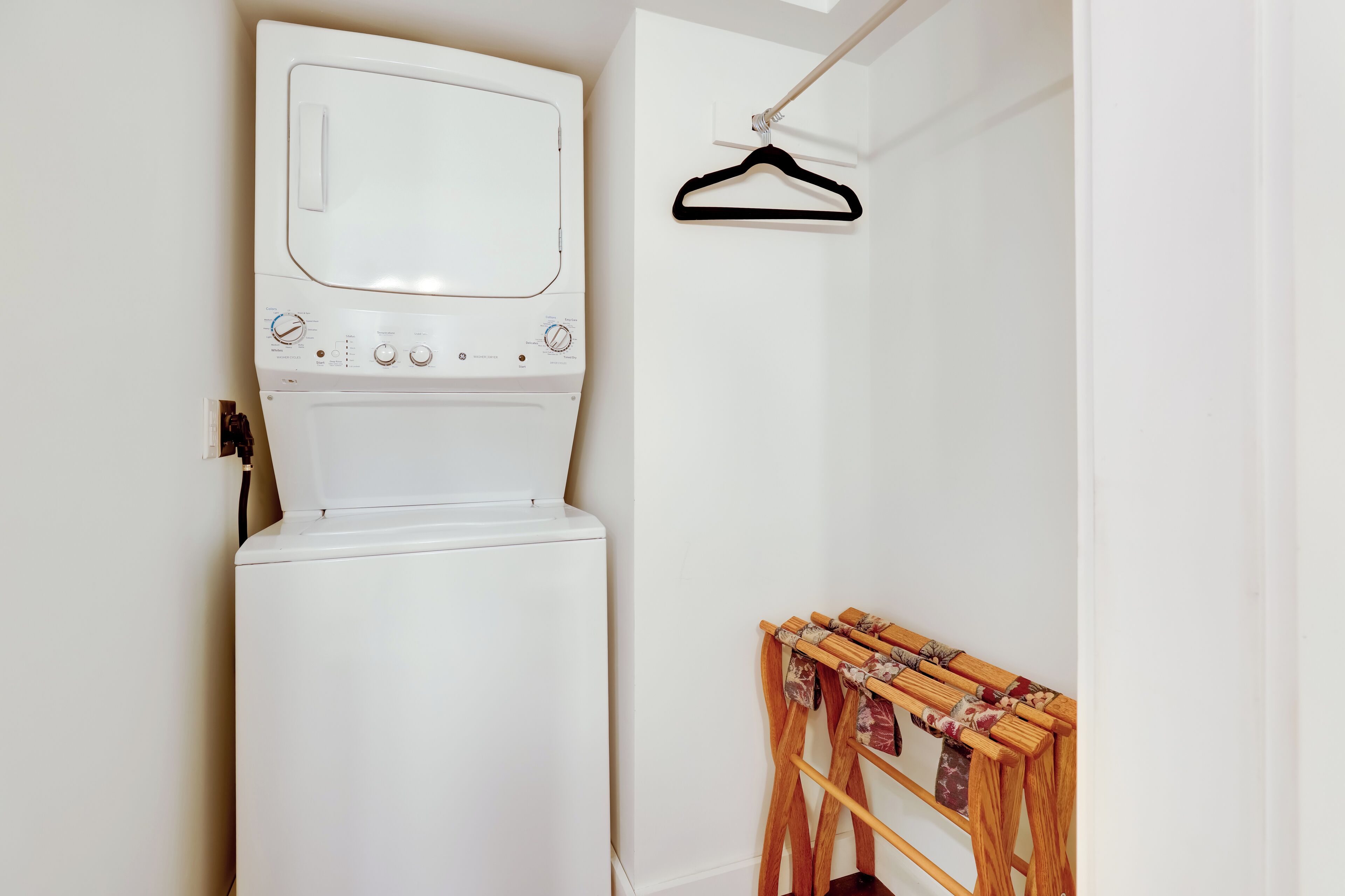 A compact laundry room with a front-loading washer and dryer, a wooden drying rack, and a black hanger on the wall.