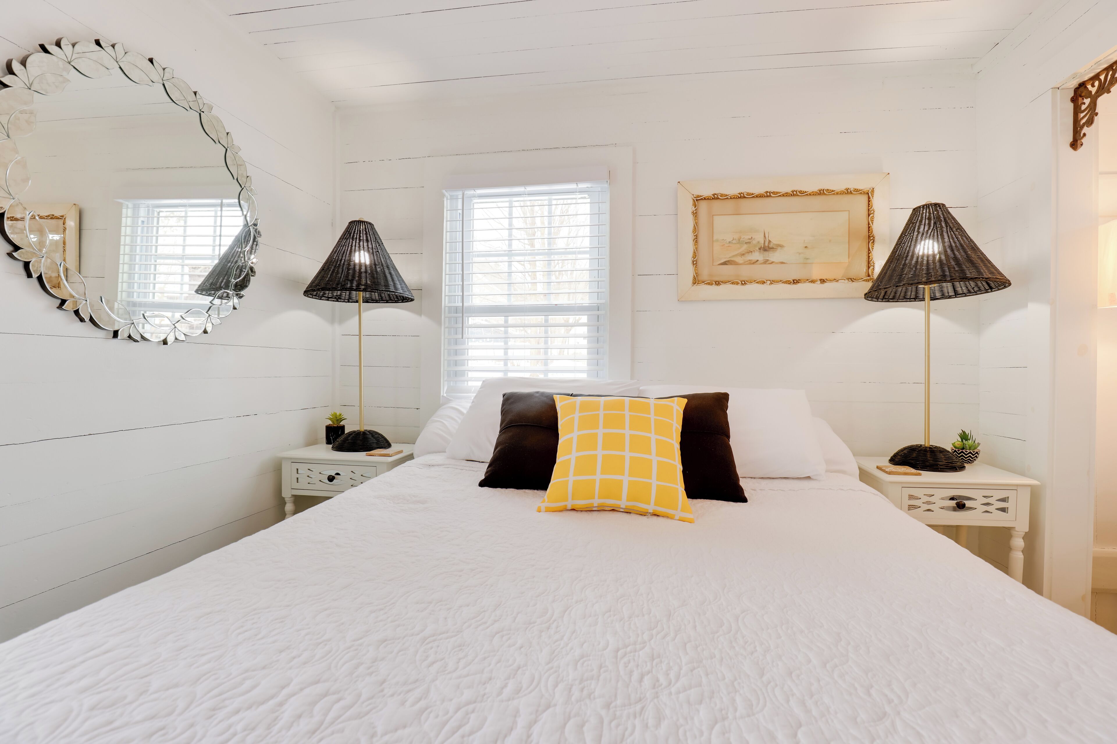 A neatly arranged bedroom with a large bed, two bedside tables, a floor lamp, a mirror, and a window with blinds.