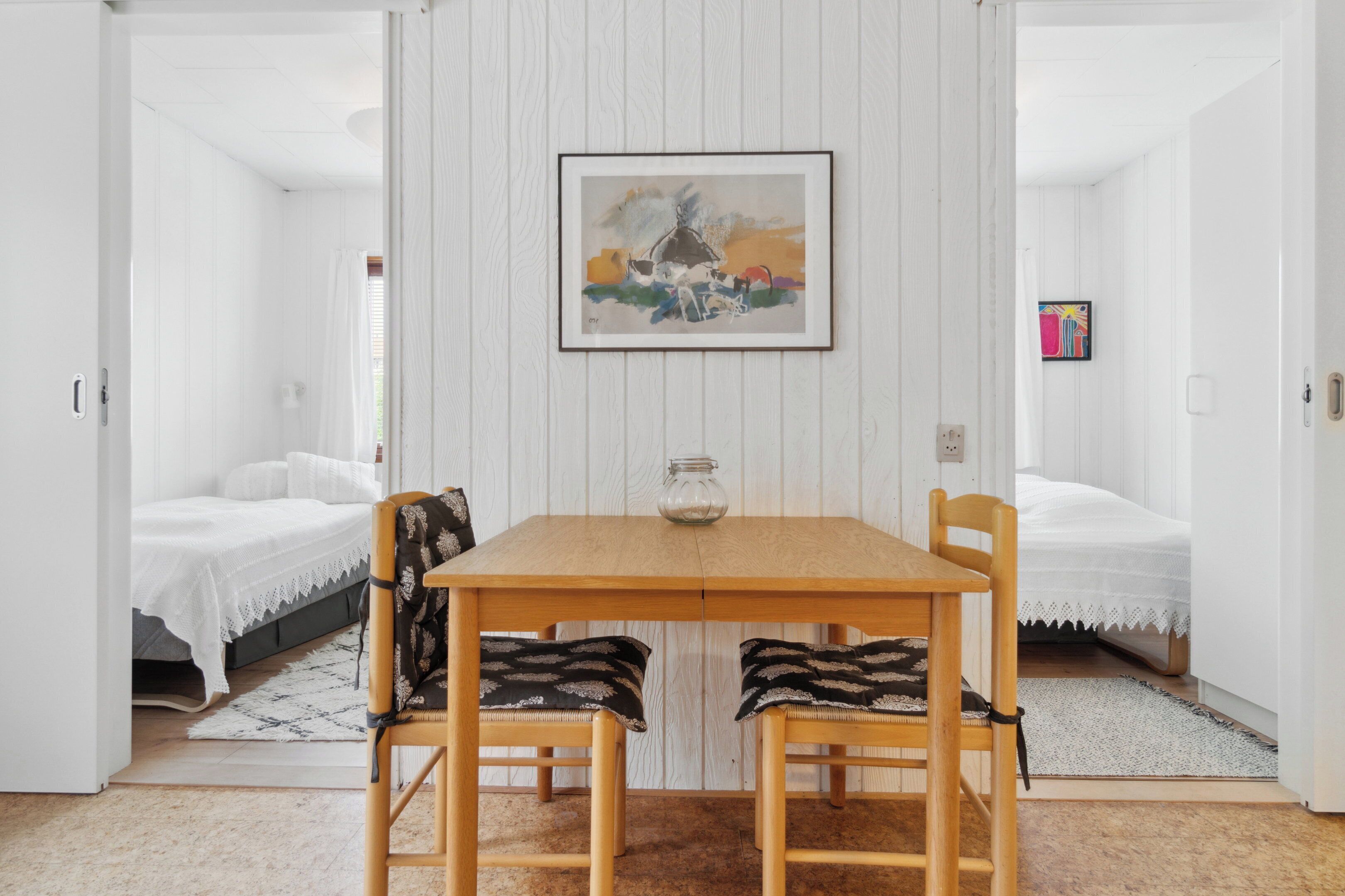 A room with a wooden table, two chairs, a bed with white bedding, and a framed painting on the wall.