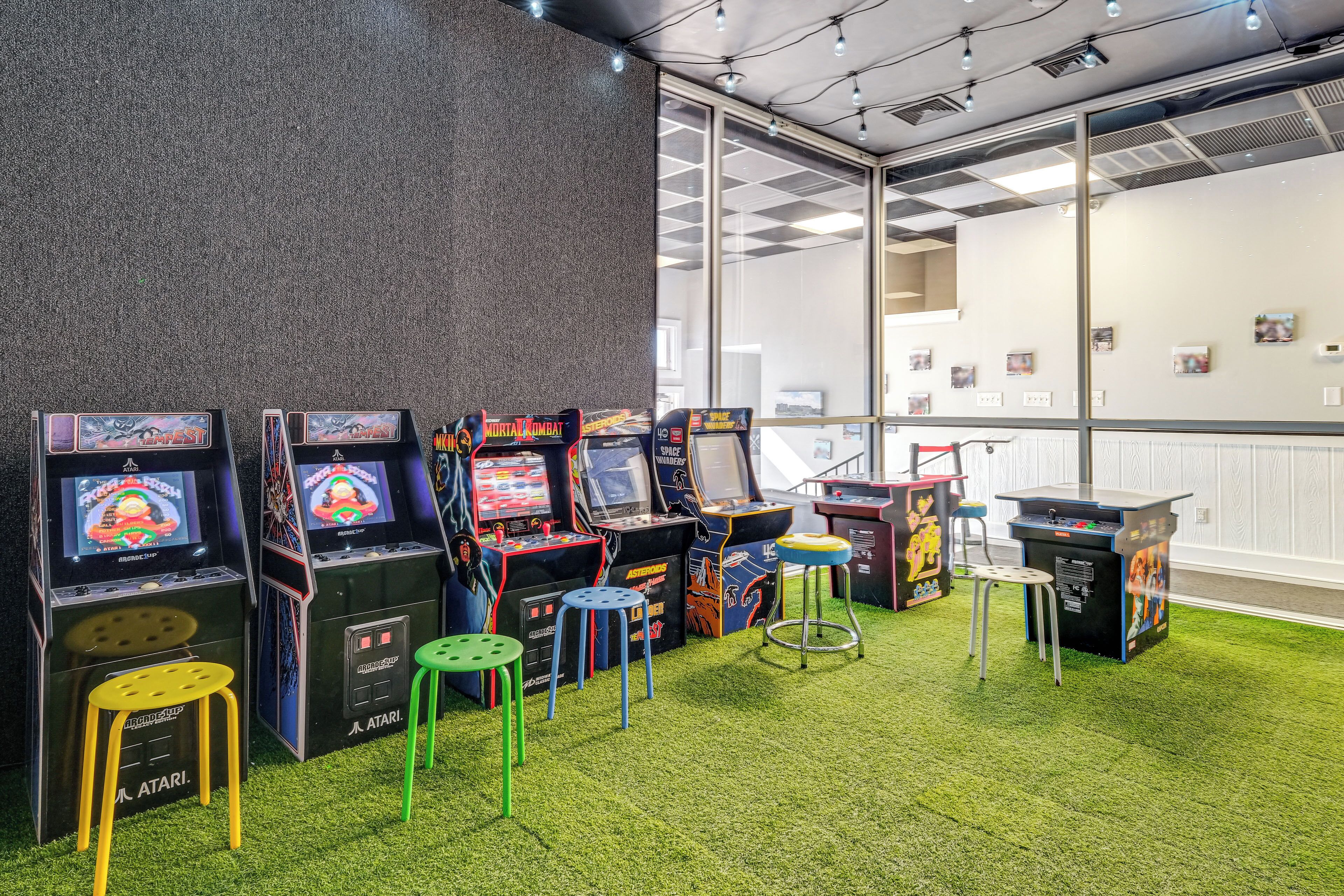 Een rij speelautomaten met krukken in een ruimte met een groen tapijt en een raam met jaloezieën.