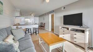 Ein Wohnzimmer mit grauem Sofa, einem Holztisch und einem Fernseher auf einer Holzkommode. Im Hintergrund ist eine Küche mit Essbereich zu sehen.