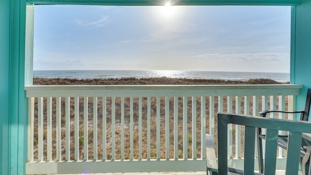 Blick vom Balkon mit weißer Reling auf einen Strand mit trockenem Gras und dem Meer.
