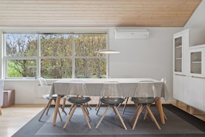 A modern dining area with a long table, four chairs, and a large window with a view of trees. - Lovely holiday home in Pøt Strandby (Juelsminde)