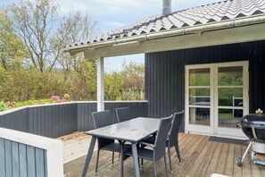 A deck with a table and chairs, a grill, and a covered area with a view of trees and a building. - Lovely holiday home in Pøt Strandby (Juelsminde)