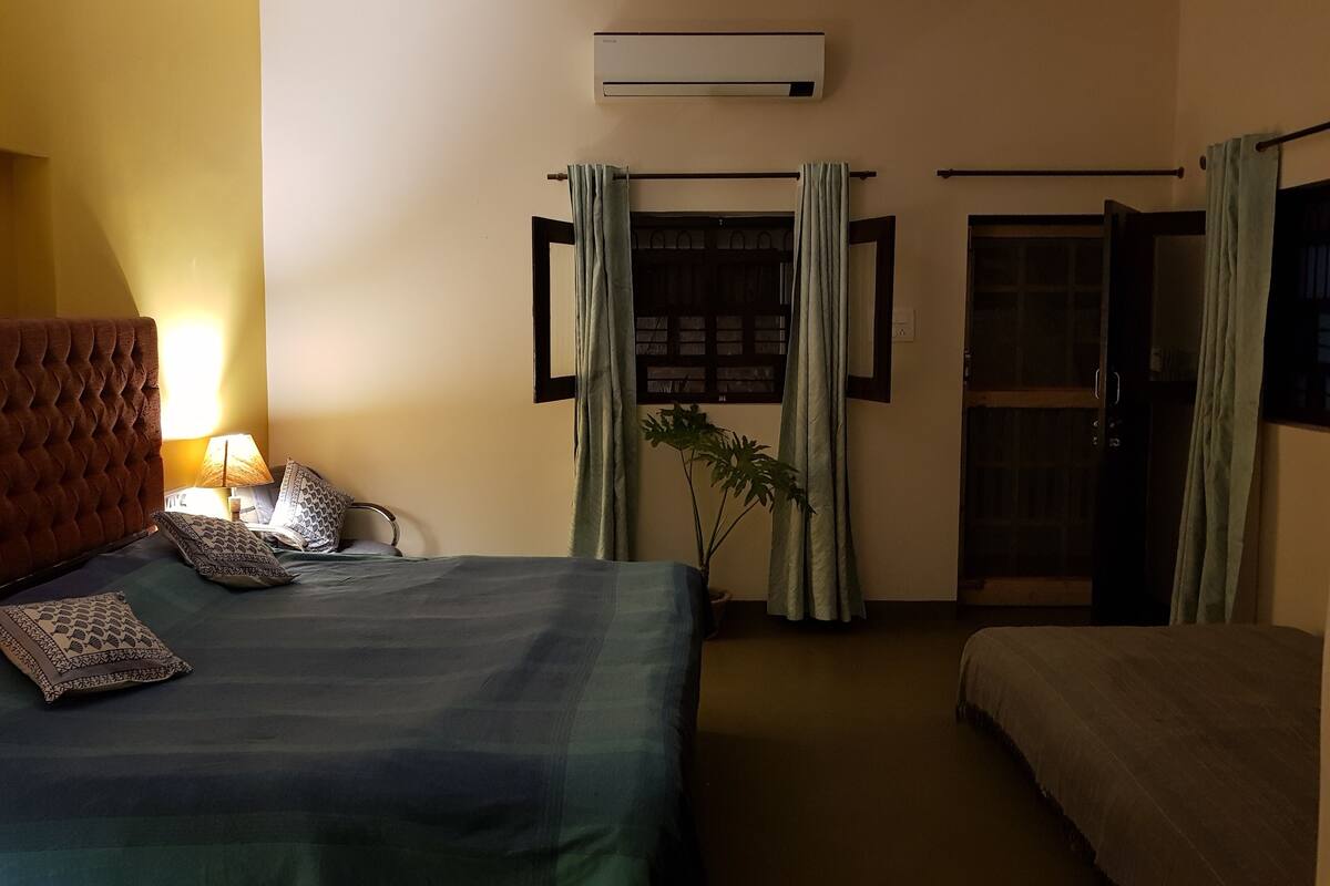 A hotel room with a bed, a bedside table with a lamp, a potted plant, and a window with curtains.