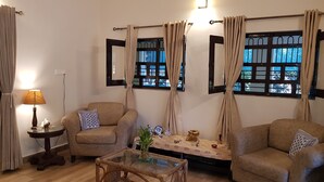 A living room with two sofas, a glass coffee table, a small side table, and a lamp. There are curtains on the windows, and a mirror is on the wall. - Koko's Farm (Mohanlalganj)