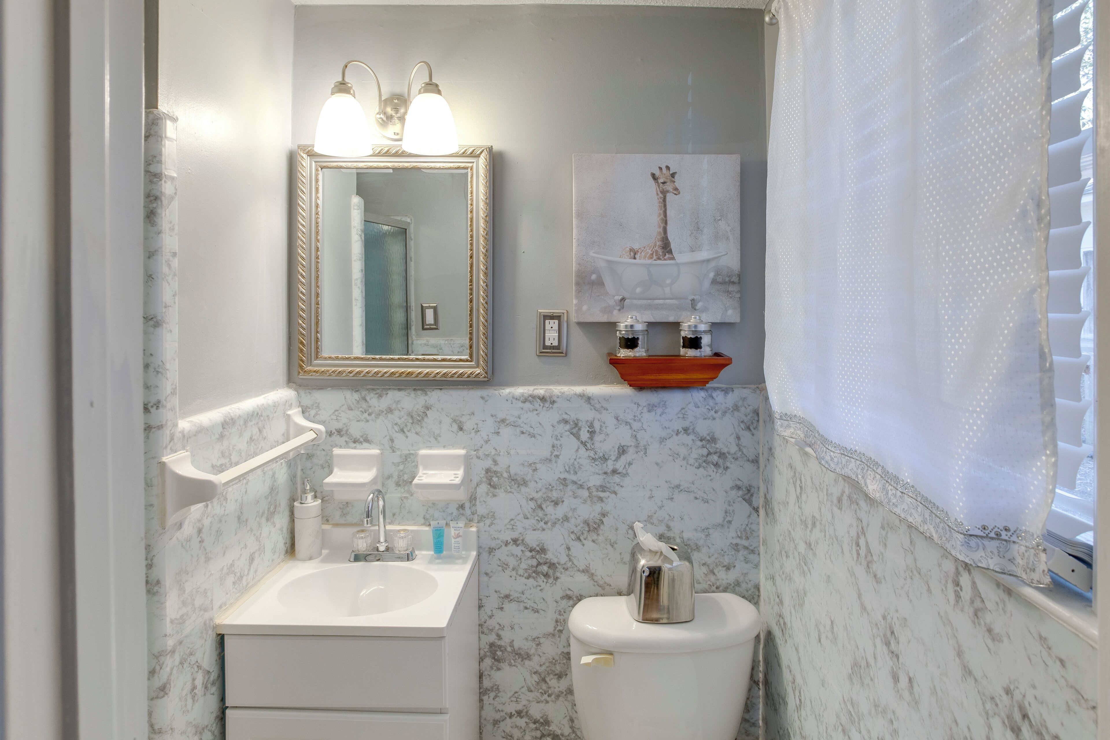 A bathroom with a marble wall, a white sink, a toilet, a mirror with a gold frame, and a decorative shelf with two bottles.