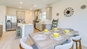 A modern kitchen with a central island, stainless steel appliances, and a dining area with a wooden table and yellow bowls. - Family-friendly Home w/ Yard in Hockley! (Hockley)