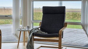 A modern room with a large window, a wooden floor, a grey upholstered chair, and a small round table with a candle. - Cozy holiday home on Orø (Orø)