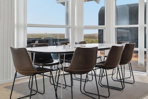 Spiseområde med et rundt bord og seks stole, store vinduer med udsigt over landskabet, og moderne arkitektur.