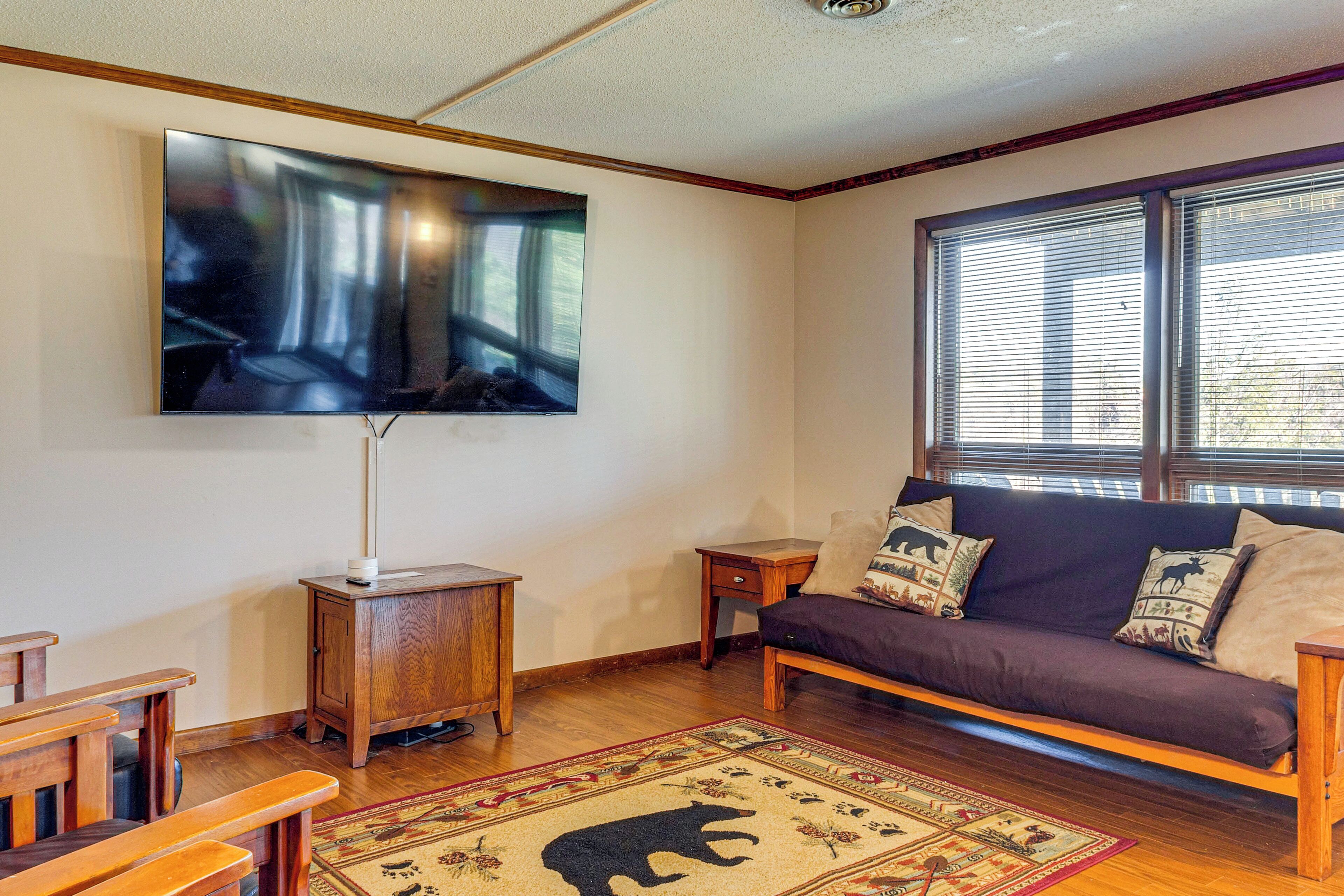Una sala de estar con un televisor de pantalla plana grande montado en la pared, un sofá de madera con cojines estampados, una mesita de madera y una alfombra decorativa con motivos de animales.