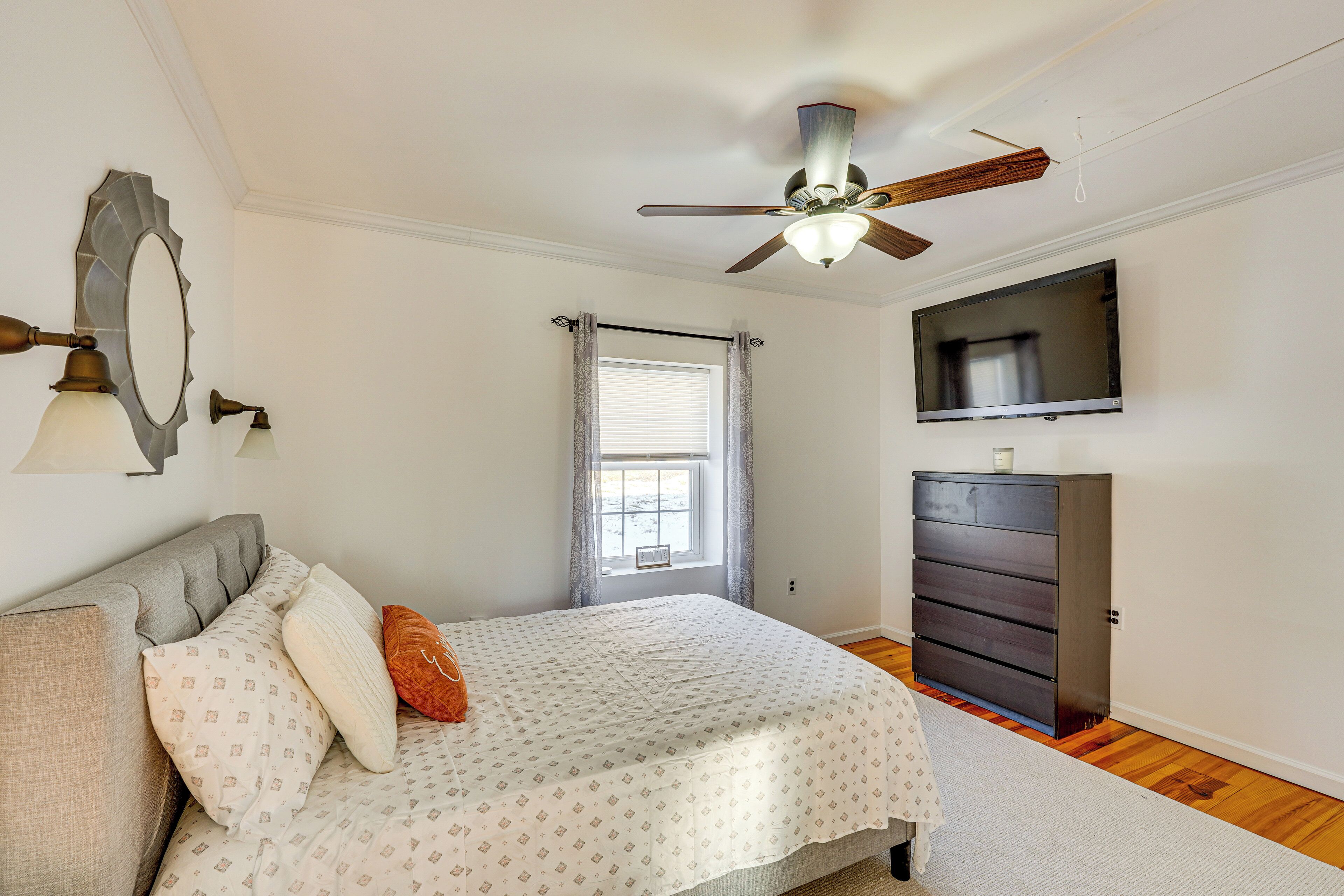 Ein Schlafzimmer mit Bett, Kommode, Deckenventilator, Fernseher und Fenster mit Vorhängen.